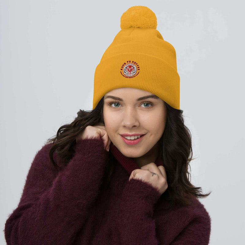 Yellow pom-pom beanie with embroidered logo worn by smiling woman in burgundy sweater